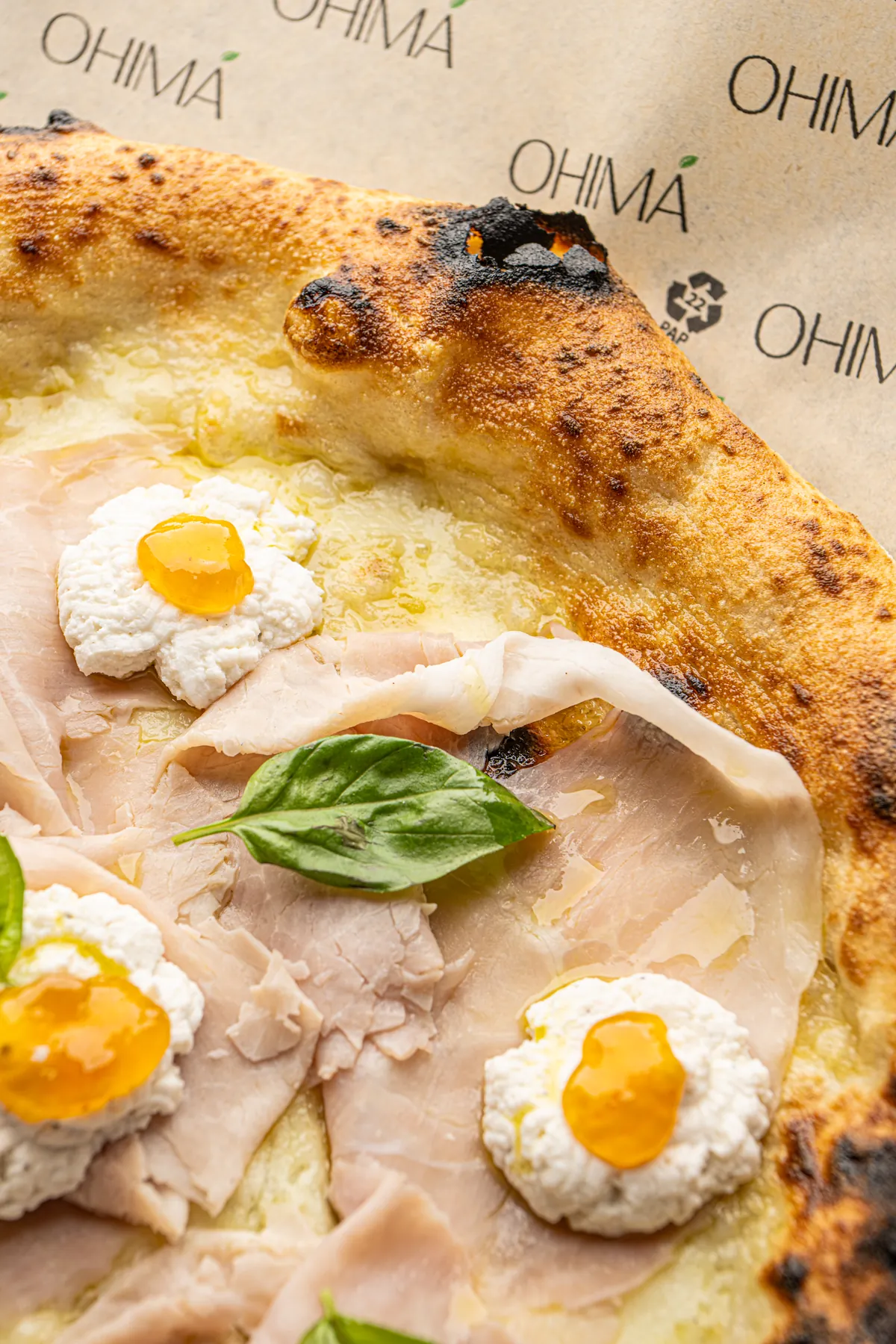 Dettaglio della pizza sulla carta con il logo Ohimà: cornicione dorato, fette di prosciutto, ricotta con una goccia di salsa arancione e basilico