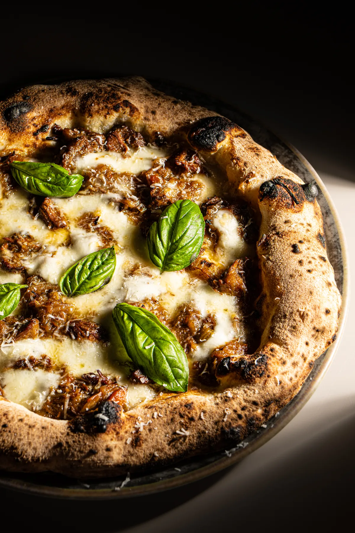 Pizza con ragù sfilacciato, mozzarella e foglie di basilico, in luce radente su fondo scuro
