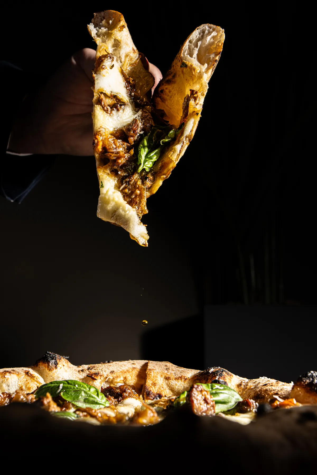 Uno spicchio di pizza tenuto in mano sopra una pizza intera: ragù, mozzarella e basilico, con una goccia d'olio che cade